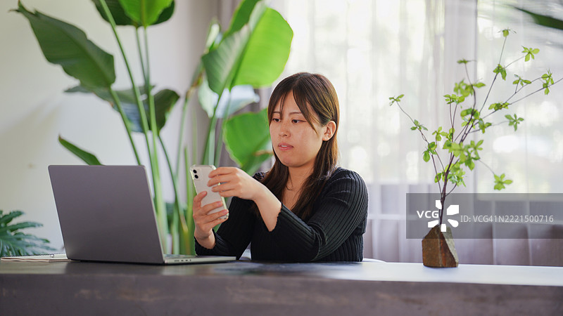 女商人在现代绿色办公室的桌子前坐着，使用笔记本电脑和智能手机工作。图片素材