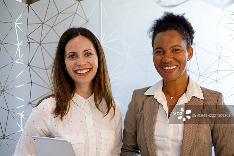 两位微笑的女商人在现代办公室中合影，其中一人手持平板电脑。图片素材