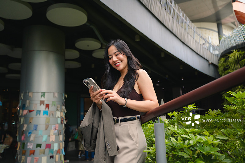 年轻的亚洲女商人在现代城市环境中微笑着使用智能手机图片素材