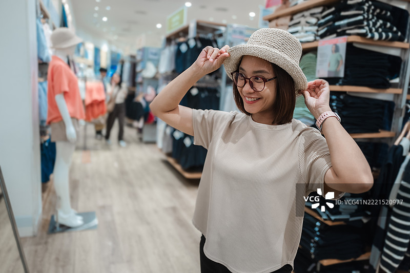 微笑的亚洲女性在时尚服装店试戴一顶时尚帽子，享受购物体验并探索时尚选择图片素材
