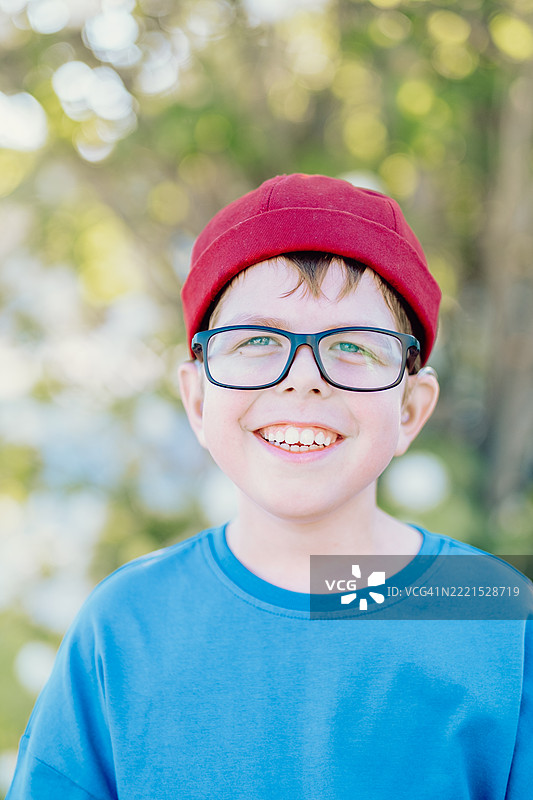 一位11岁男孩的明亮肖像，戴着助听器和眼镜，穿着红色夹克，露出洁白整齐的牙齿微笑。图片素材
