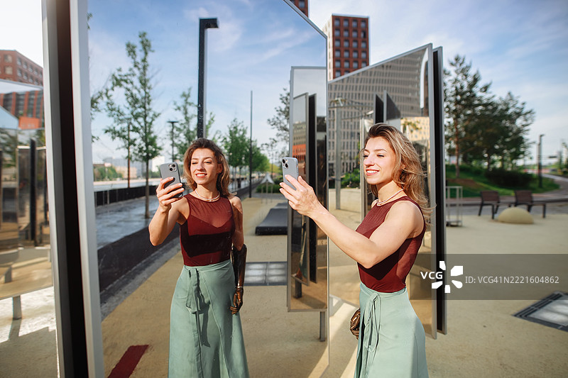 穿着夏季裙子的苗条女性在镜子前排练舞蹈，创造出自信与快乐的魔幻时刻，并用智能手机录制自己。图片素材