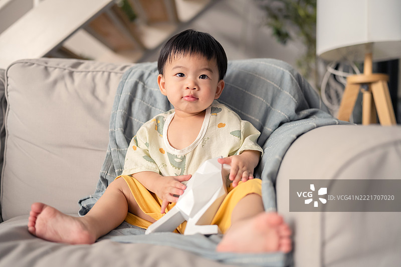 可爱的亚洲小男孩悠闲地坐在简约风格的布艺沙发上，开心地微笑着，独立走动并倚靠在沙发上，捕捉了早期儿童发展、舒适和家庭生活方式的瞬间。图片素材