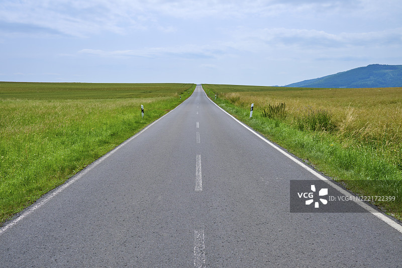 一条笔直的乡村道路穿过宽阔的田野，通向远处的山丘，天空布满云彩，位于德国黑森州的巴德苏登-阿伦多夫镇，韦拉-迈斯纳县。图片素材