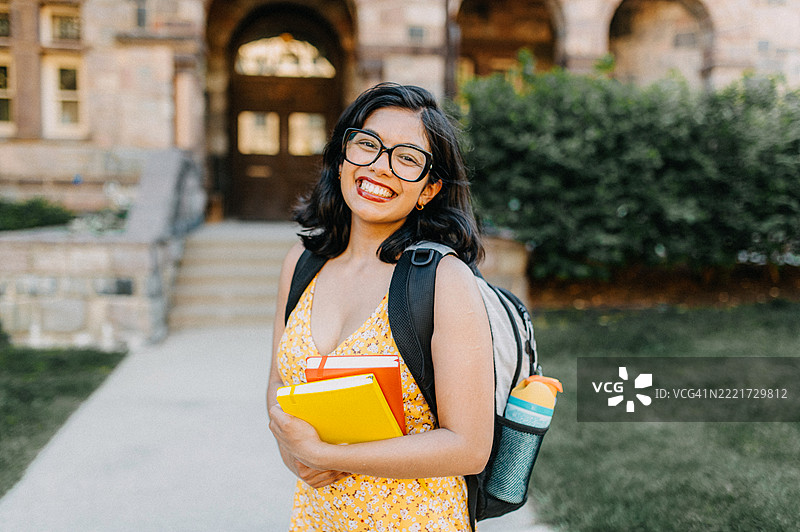一位戴眼镜的年轻女性自信地微笑,手里拿着五颜六色的书籍,背着背包在户外。图片素材