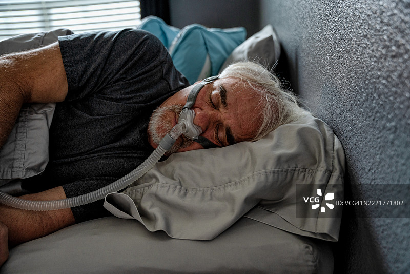 一位60岁的男性在床上使用CPAP机睡觉。该机器帮助他更轻松地呼吸，并防止睡眠呼吸暂停，这是一种常见的睡眠障碍。图片素材