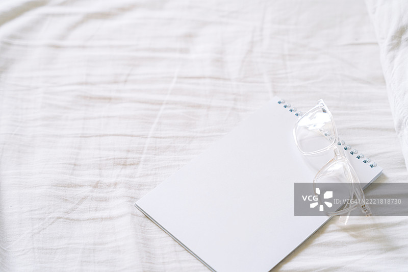 空白页笔记本和清晰眼镜放在皱褶的白色床单上，阳光洒下。柔和的色调和简单的构图反映出宁静的专注、反思和舒适的高效。图片素材