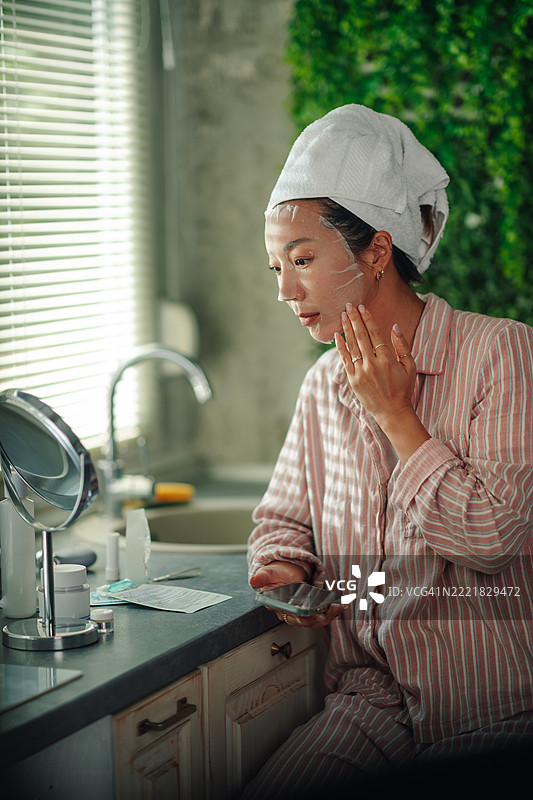 年轻的亚洲女性一边阅读智能手机上的说明，一边涂抹化妆面膜图片素材