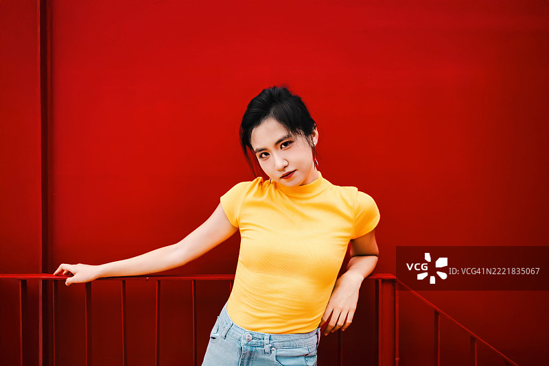 自信的年轻女性穿着黄色T恤，倚靠在红色墙壁上，展现都市态度和现代街头风格。图片素材