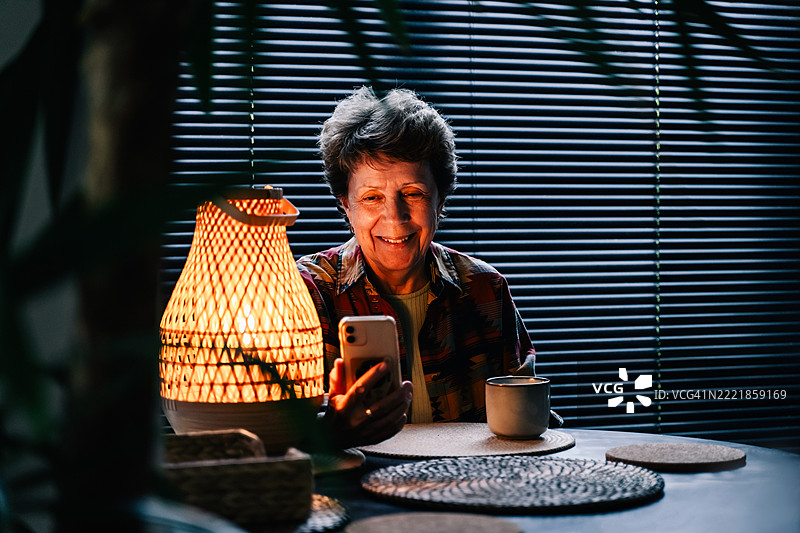 穿着色彩鲜艳衬衫的老年女性在夜晚使用智能手机，旁边有一个发光的藤编灯，享受着温暖的饮品，营造出舒适而富有情调的氛围。图片素材