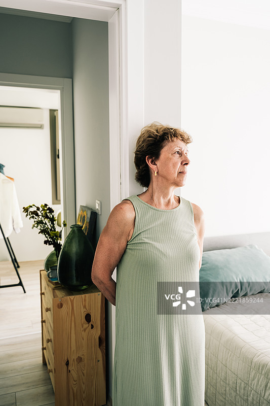 一位成熟的女性站在卧室里，传达出情感或沉思的状态。退休和孤独的概念。图片素材