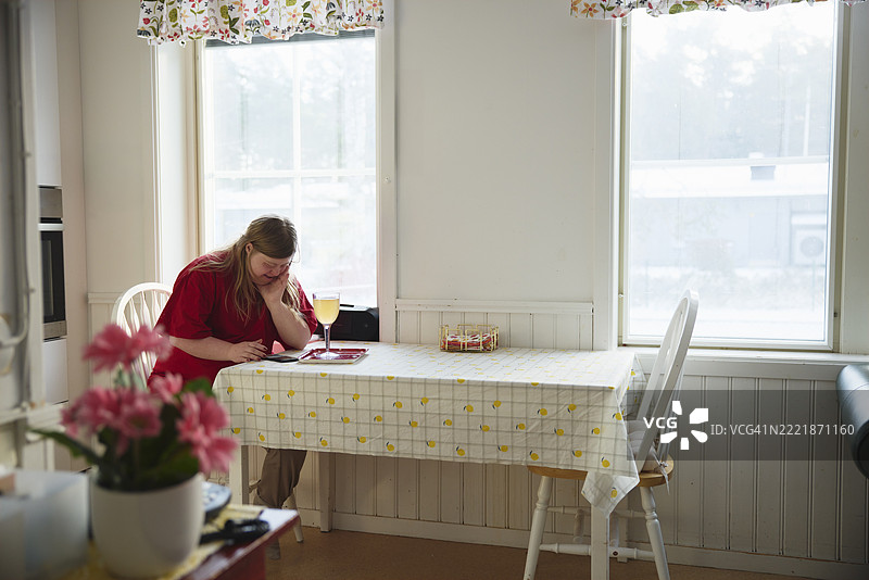坐在家中厨房窗边的桌子旁，患有唐氏综合症的女性正在使用智能手机。图片素材