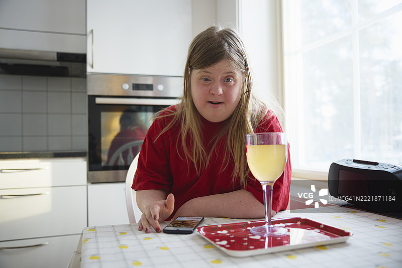 在家厨房里，坐在桌子旁的唐氏综合症女性，手里拿着一杯果汁。图片素材