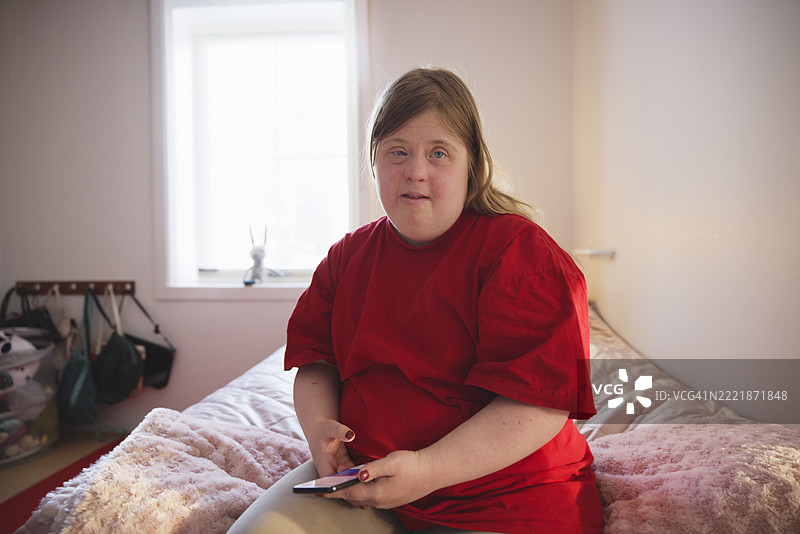 在养老院的卧室里，坐在床上的唐氏综合症女性手持智能手机的肖像。图片素材