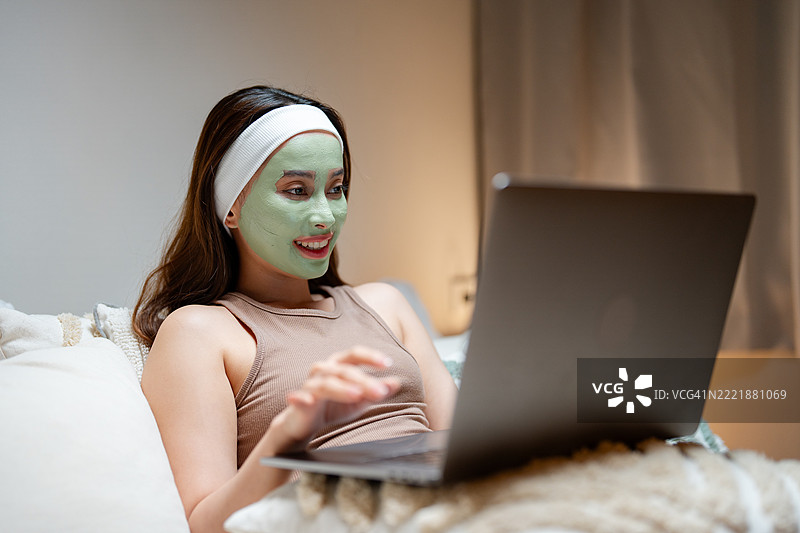 微笑的年轻女性坐在床上，戴着有机面膜，一边使用笔记本电脑。图片素材