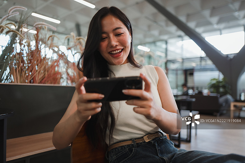 年轻的亚洲办公室工作人员在休息时玩手机游戏，微笑着在轻松的环境中享受乐趣。图片素材
