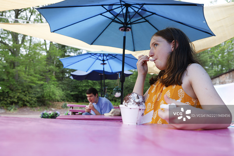 与自闭症兄弟的冰淇淋休息时间。一个青春期女孩和一个少年在蓝色伞下共享户外甜点。图片素材