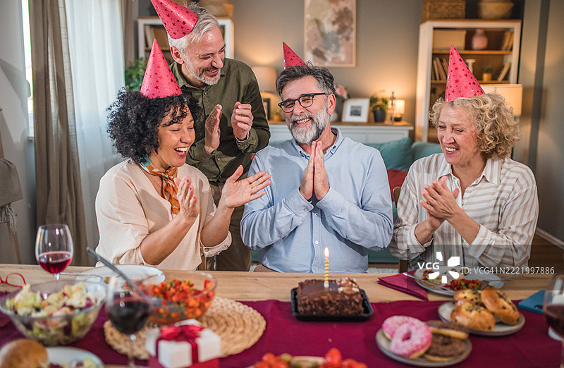 一位无忧无虑的老年人与朋友们一起享受生日派对。他们为他唱生日歌，等待他吹灭生日蛋糕上的蜡烛。图片素材