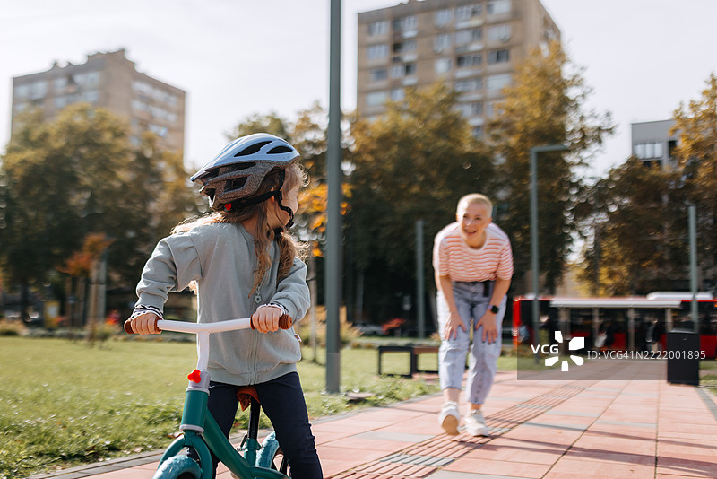 小女孩戴着自行车头盔在公园骑自行车，母亲在她身后。图片素材