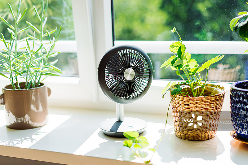 现代桌面风扇放在窗台上，周围环绕着盆栽绿植图片素材