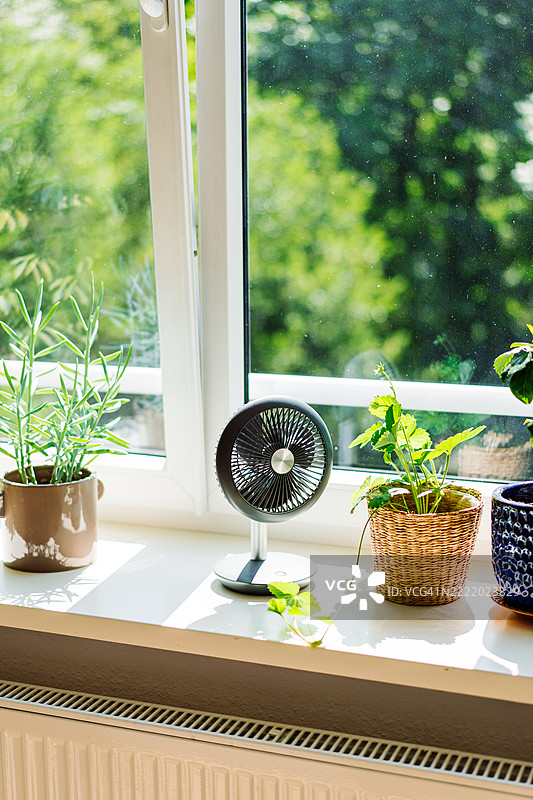 现代桌面风扇放在窗台上，周围环绕着盆栽绿植图片素材