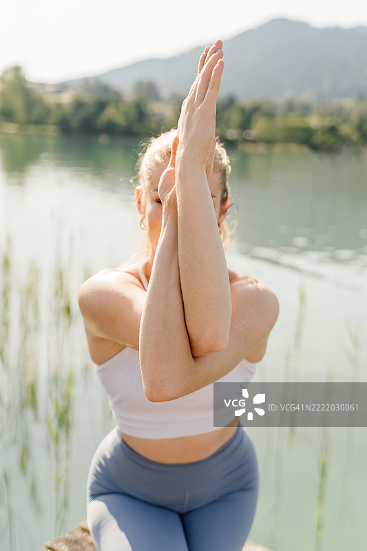 年轻女性在湖边的木平台上做早晨练习，摆出加鲁达姿势，背景是山脉。图片素材