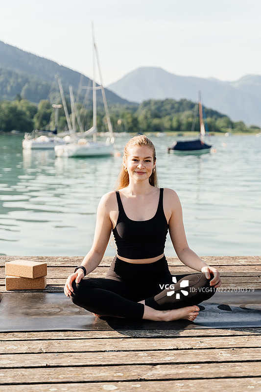 穿着瑜伽服的女性坐在湖边木栈道上以莲花坐姿冥想图片素材