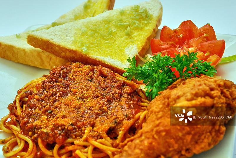 意大利面上覆盖着浓郁的肉酱，配有一块炸鸡和两片蒜蓉面包。图片素材