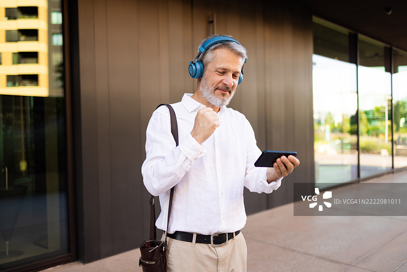 快乐的中年商人戴着耳机听音乐，并在智能手机上庆祝成功。图片素材