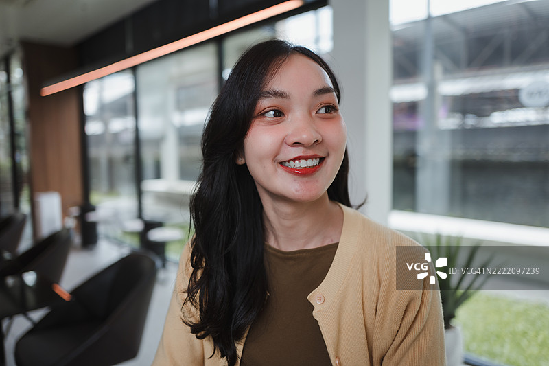 一位年轻的亚洲女性在明亮现代的办公室里微笑着望向远方，散发着积极和自信的气息。图片素材