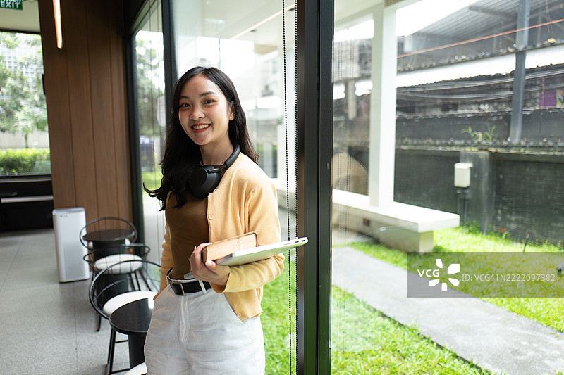 微笑的年轻亚洲学生在现代大学图书馆里手持一本书和一台笔记本电脑的肖像图片素材