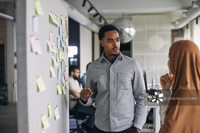 年轻男性商务专业人士在办公室的公告板旁与戴头巾的女性同事讨论图片素材