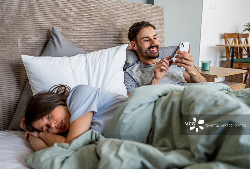 男人在床上使用智能手机，而女人在睡觉图片素材