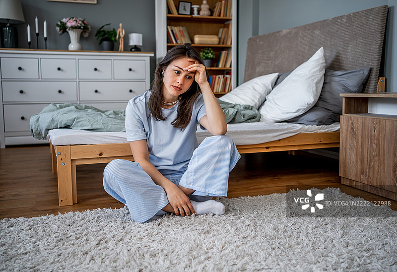伤心的年轻女性坐在卧室地板上，手触额头。图片素材
