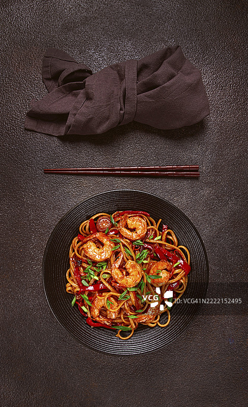 黑碗中的乌冬炒虾面，配有红椒和青葱，旁边放着筷子，背景为深色。图片素材