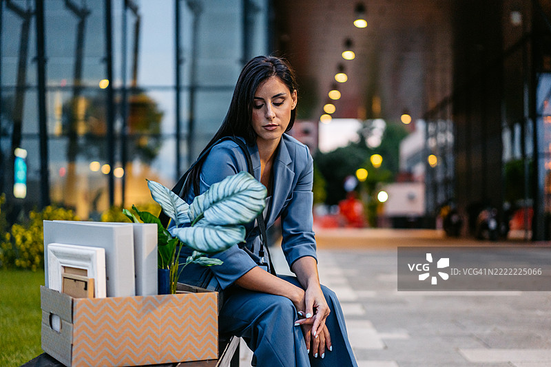 伤心的年轻女商人坐在公司大楼前的长椅上，手里拿着一箱办公用品。图片素材