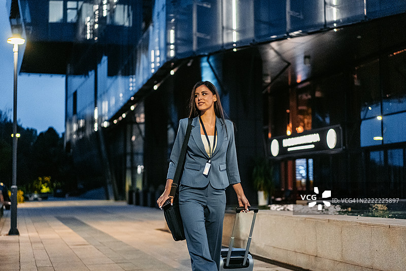 年轻女商人在夜晚走过一座企业大楼，推着行李箱。图片素材