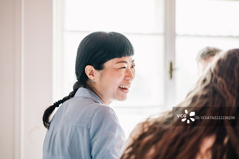 一位穿着蓝色衬衫的微笑亚洲女性特写，柔和的自然光照亮她在办公室交谈时的笑容。图片素材