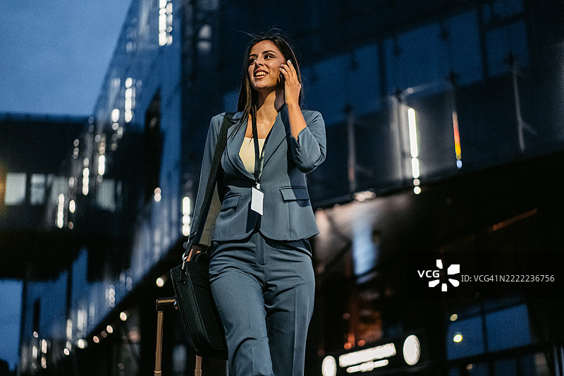 年轻女商人在夜晚的公司大楼前一边打电话一边拖着行李箱图片素材