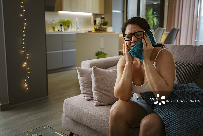 一位女性坐在沙发上，因严重牙痛而在下巴上敷冰袋。她感到不适和不满，闭着眼睛待在家中。图片素材
