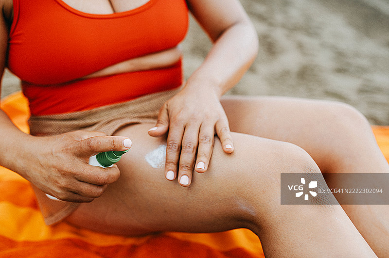一位穿着红色泳衣的女性坐在海滩上的橙色毛巾上,正在给大腿涂抹防晒霜。图片素材