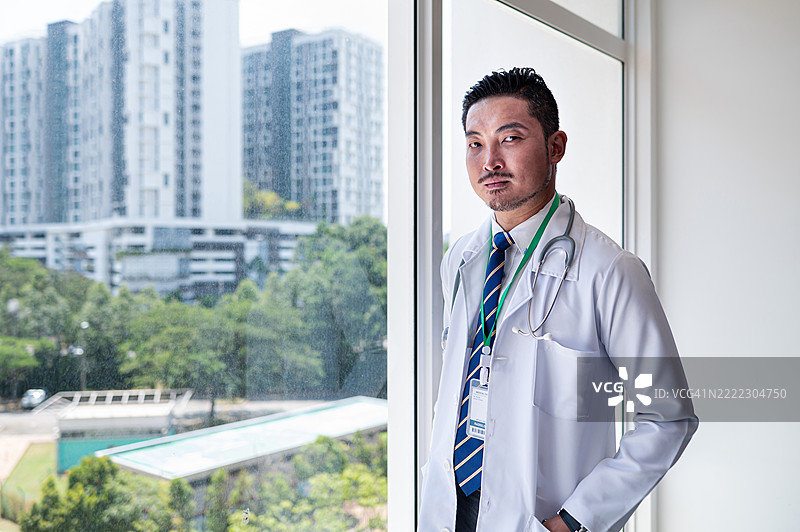 中年男性医生穿着实验室制服，面向镜头，站在办公室窗边。图片素材