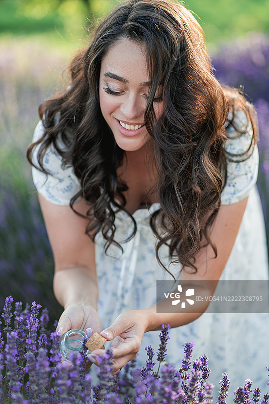 年轻美丽的女人在田野里采摘薰衣草花。手中拿着一瓶精油。图片素材