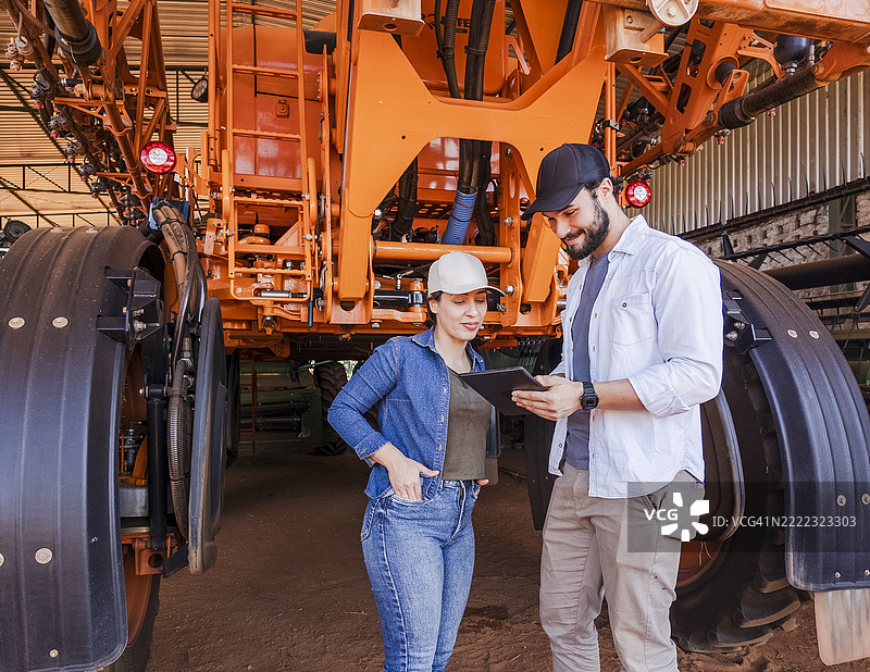 农业专家团队在乡村棚屋旁使用平板电脑，旁边是农业机械。图片素材