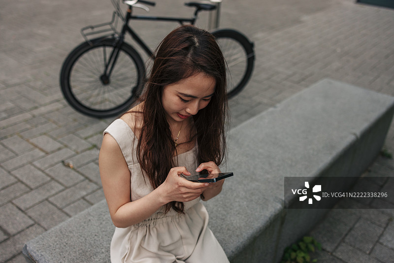 年轻女性坐在长椅上，使用智能手机，旁边放着她的自行车。图片素材