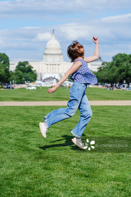 年轻女性在绿草地上充满活力地跳跃，背景是美国国会大厦。在美国国会大厦前的快乐跳跃捕捉了能量和自由。华盛顿，哥伦比亚特区，美国。图片素材