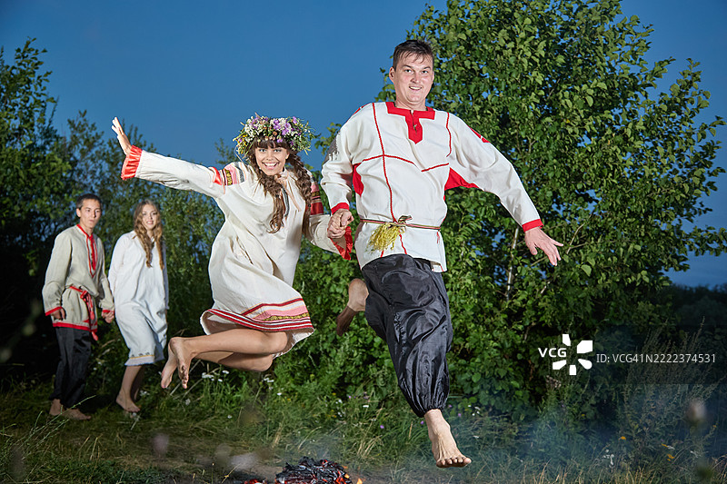 年轻漂亮的女人和她的父亲穿着民俗服装，手牵手跳过篝火。图片素材