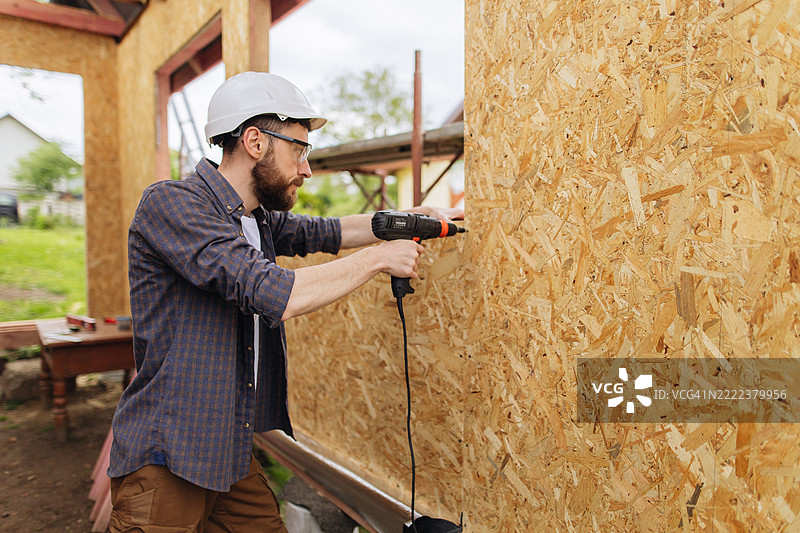 戴着安全帽的男子在建造房屋框架时用螺丝刀固定OSB板。图片素材