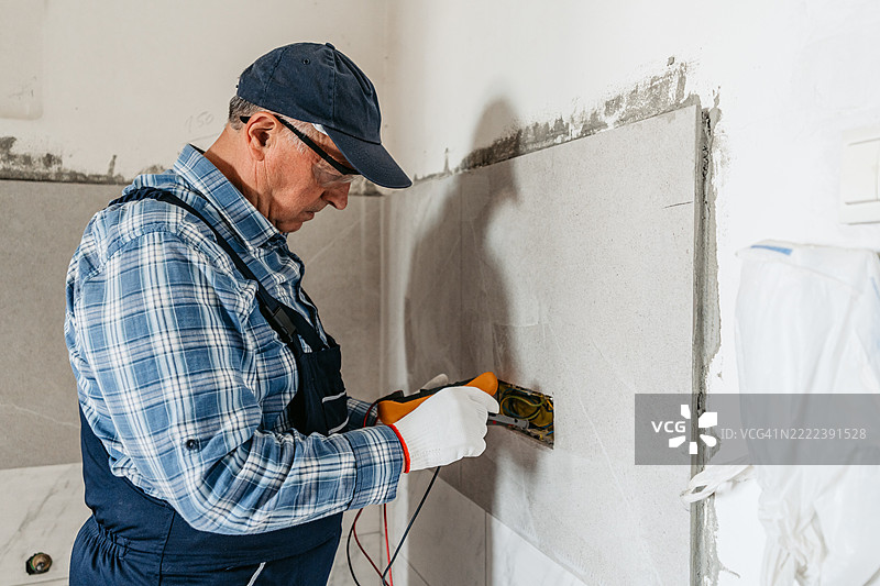 一位年长男性电工在正在装修的空公寓中使用万用表检查电缆。图片素材