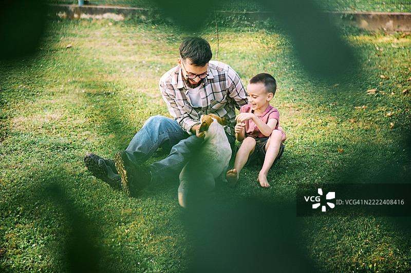 父亲和儿子坐在后院的草坪上，和他们的狗一起玩得很开心。图片素材